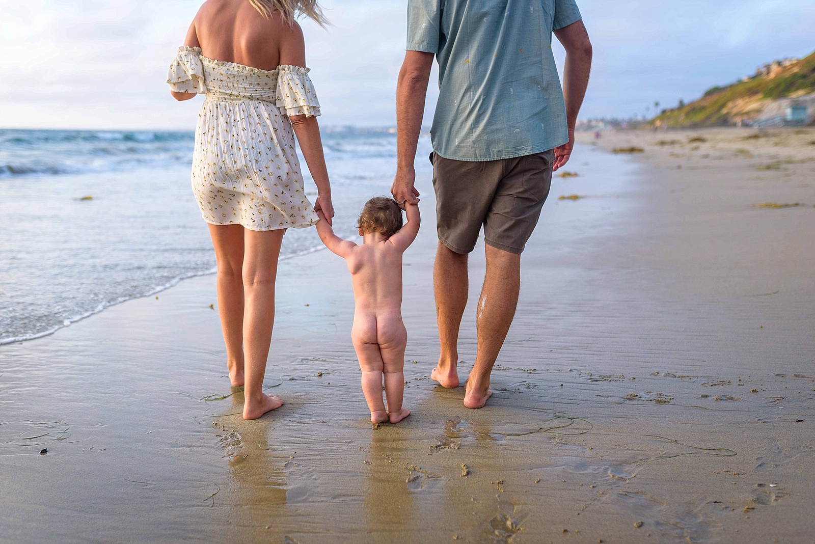 parents walk with naked toddler on the beach